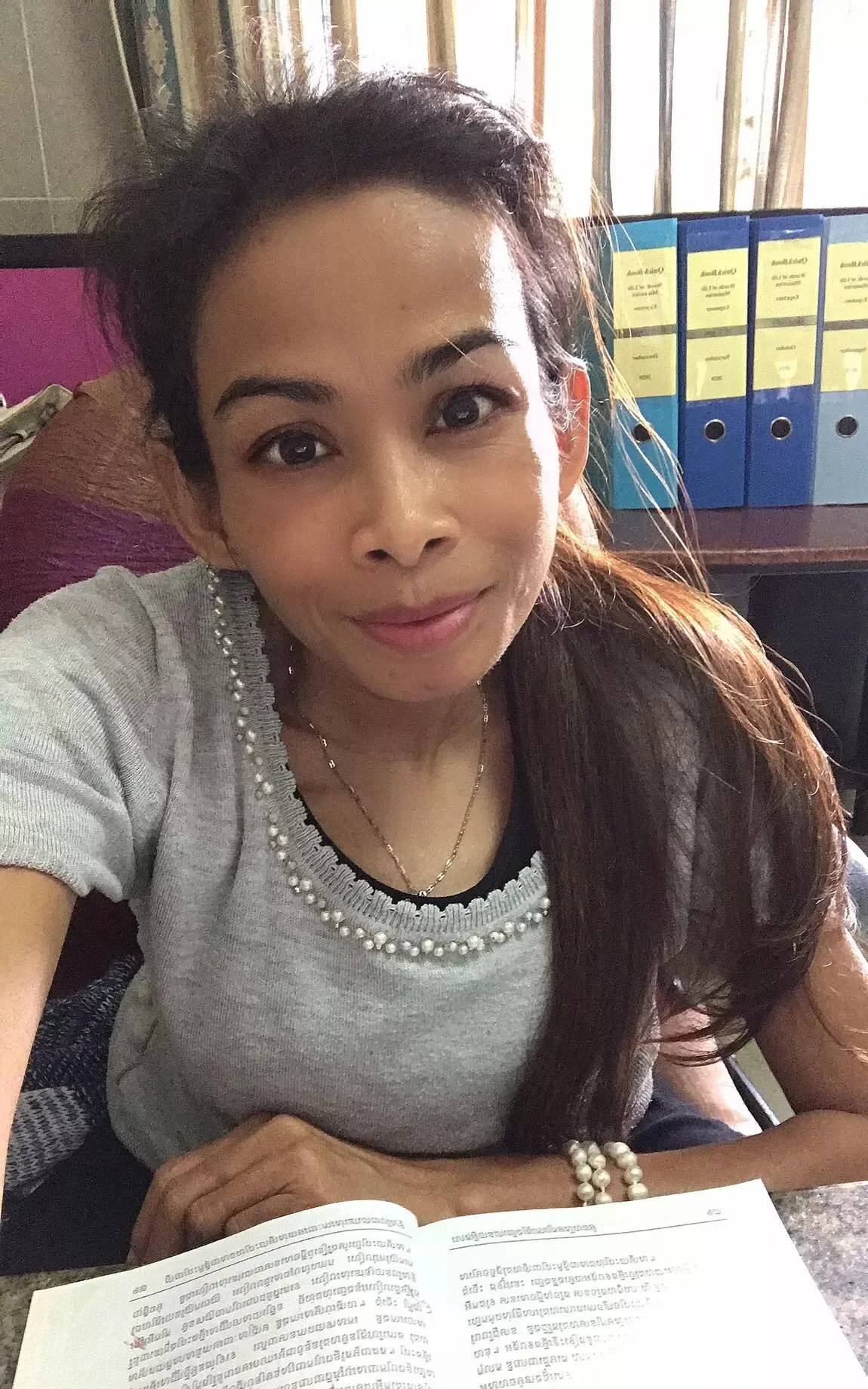 Naleng Real at her desk with an open Khmer book, ministry binders on the shelf behind her