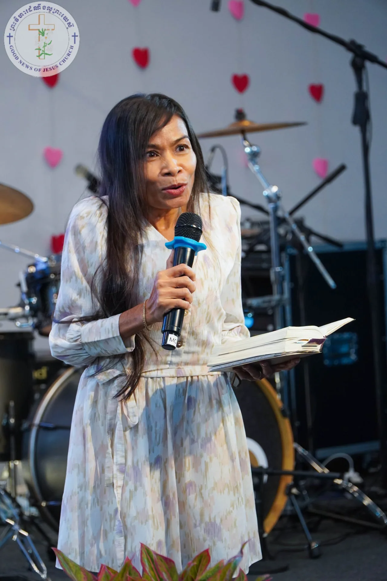 Naleng Real speaking with a microphone and an open Bible on the platform at Good News of Jesus Church
