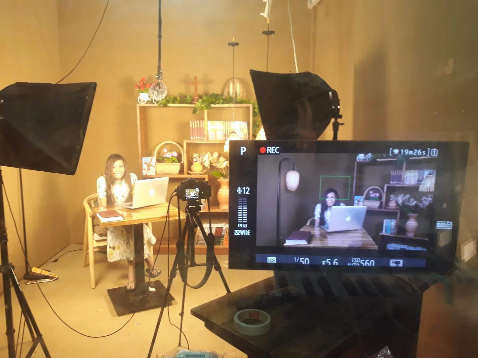 A recording set with softbox lights and a camera on a tripod, Naleng Real seated at a desk with the monitor showing the shot
